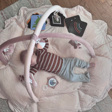 Baby lying on a play mat with a pink baby gym and toys around