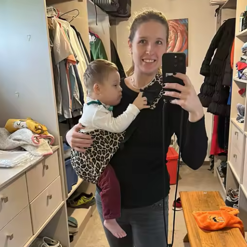 Woman holding a baby and taking a mirror selfie in a room with shelves and clothes.