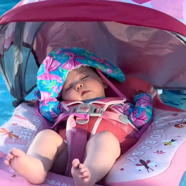Baby in a pink life jacket and hat sitting in a baby seat with a colorful canopy.