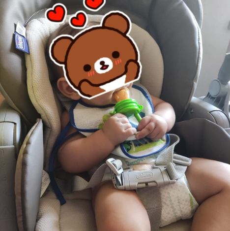 Child in a car seat with a bib and toy, wearing a bear mask filter.
